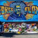 A woman holding a Black Lives Matter sign sitting before a mural of George Floyd, which is serving as a makeshift memorial.