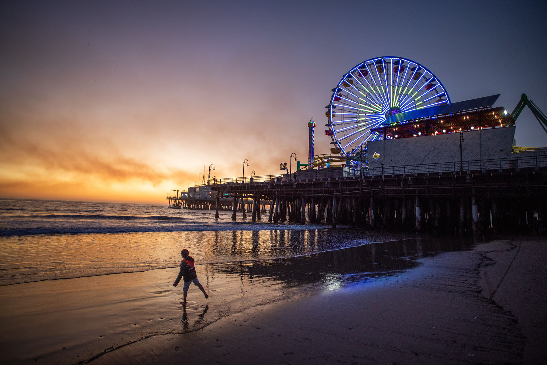 A boy plays on the beach with the Santa Monica pier on the background with smoke from the Palisades Fire seen in the sky.