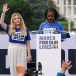 Two women stand at a podium while people in a crowd raise their hands in front of them.