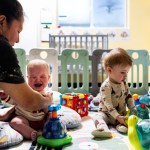 A teacher comforts a young child at a daycare as other toddlers play.