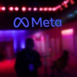 The Meta logo is seen on a glass surface with silhouettes of people and colorful purple and red lighting in the background.