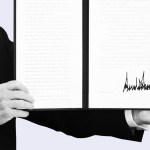 A close-up of President Trump holding open a signed executive order with a large signature visible on the right page.