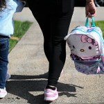 A mother holds her child's backpack and her child's hand.