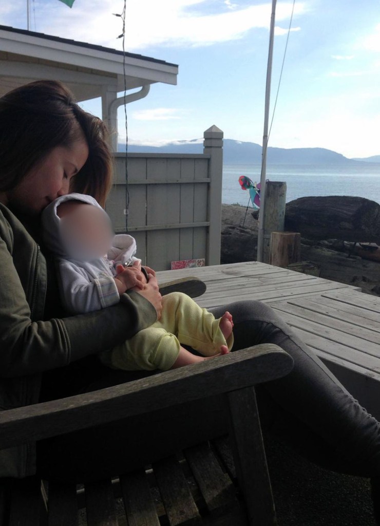 Jewel Lazaro sits outdoors on a wooden bench, cradling and kissing Lucia, whose face is blurred; behind them is a waterfront view with mountains.
